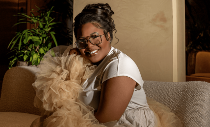Woman smiling, wearing glasses, sitting on a sofa with a fluffy dog, in a cozy indoor setting.