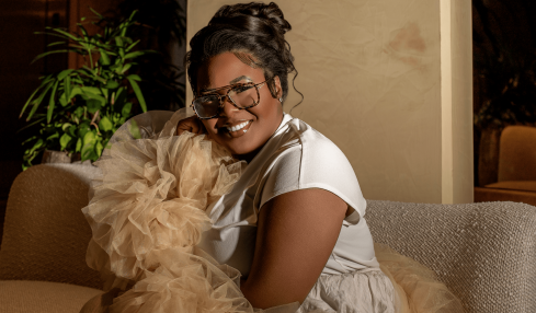 Woman smiling, wearing glasses, sitting on a sofa with a fluffy dog, in a cozy indoor setting.