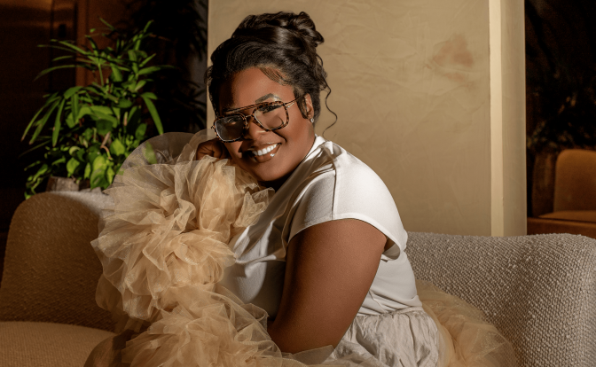 Woman smiling, wearing glasses, sitting on a sofa with a fluffy dog, in a cozy indoor setting.