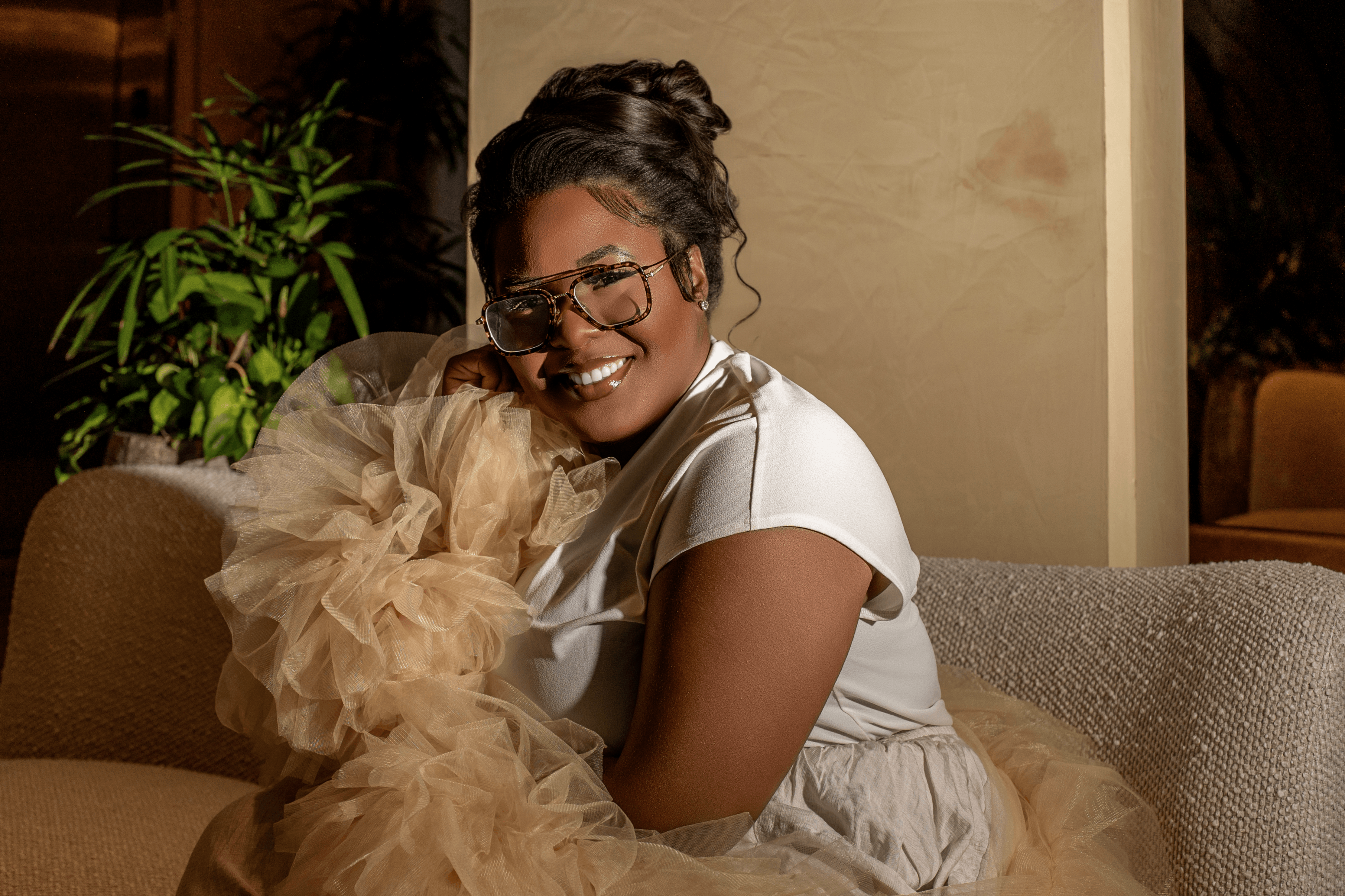 Woman smiling, wearing glasses, sitting on a sofa with a fluffy dog, in a cozy indoor setting.