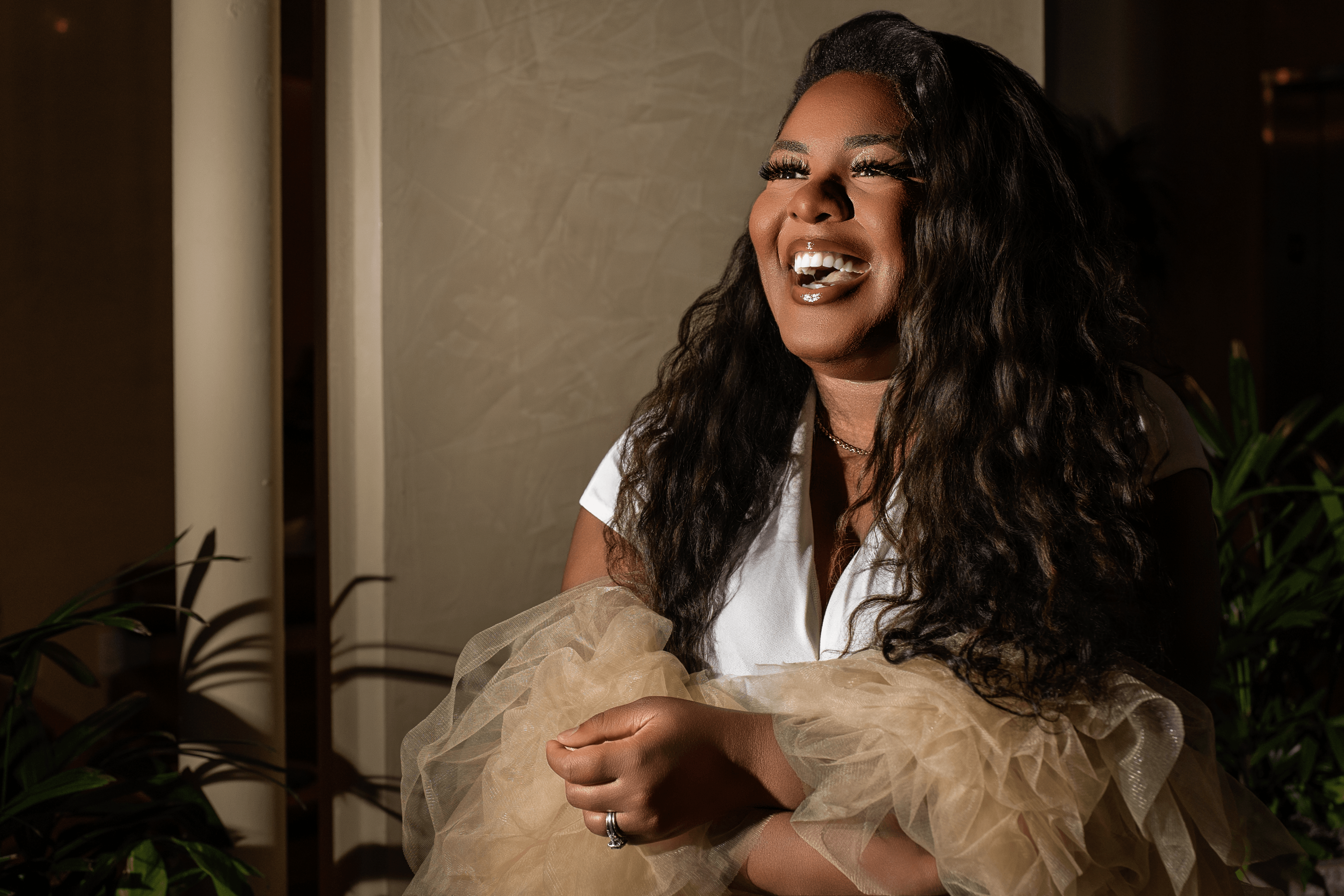 Woman with long curly hair smiling, wearing a white top and beige tulle sleeves, in a dimly lit room with plants.