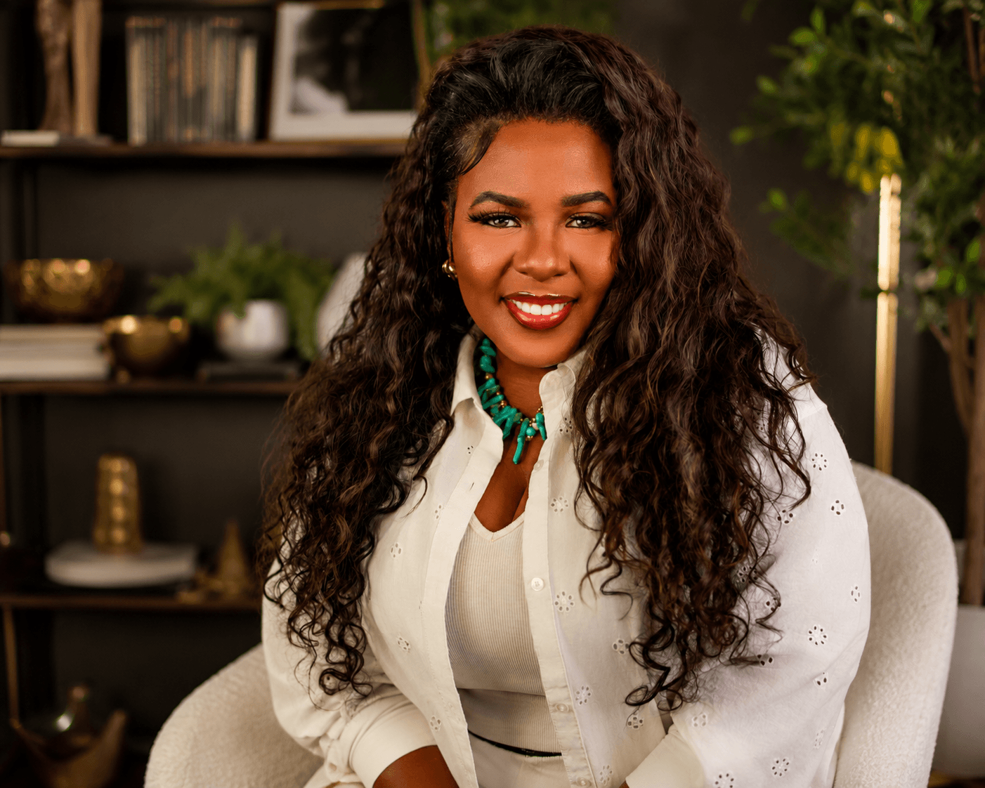 Woman with long curly hair smiling, sitting indoors with bookshelves and plants in background.